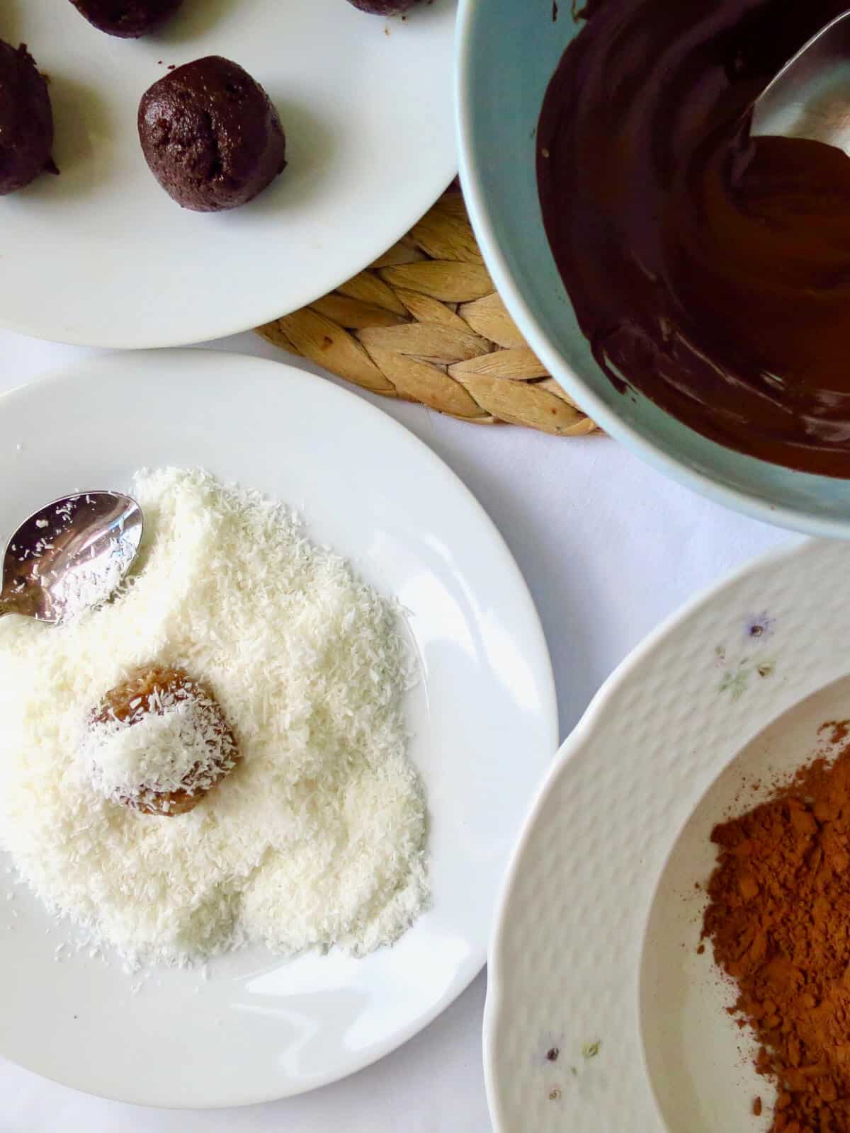 A demonstration of carrot cake ball being coated in coconut. Cocoa powder and chocolate ganache in bowls next to it.