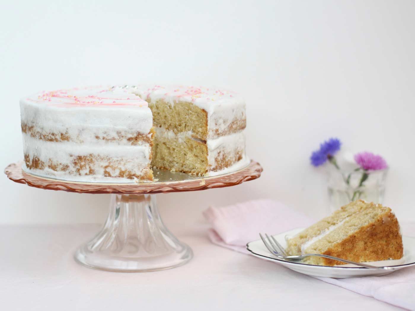 Fluffy gluten dairy free vanilla cake on cake stand with one slice of cake on a plate next to it.