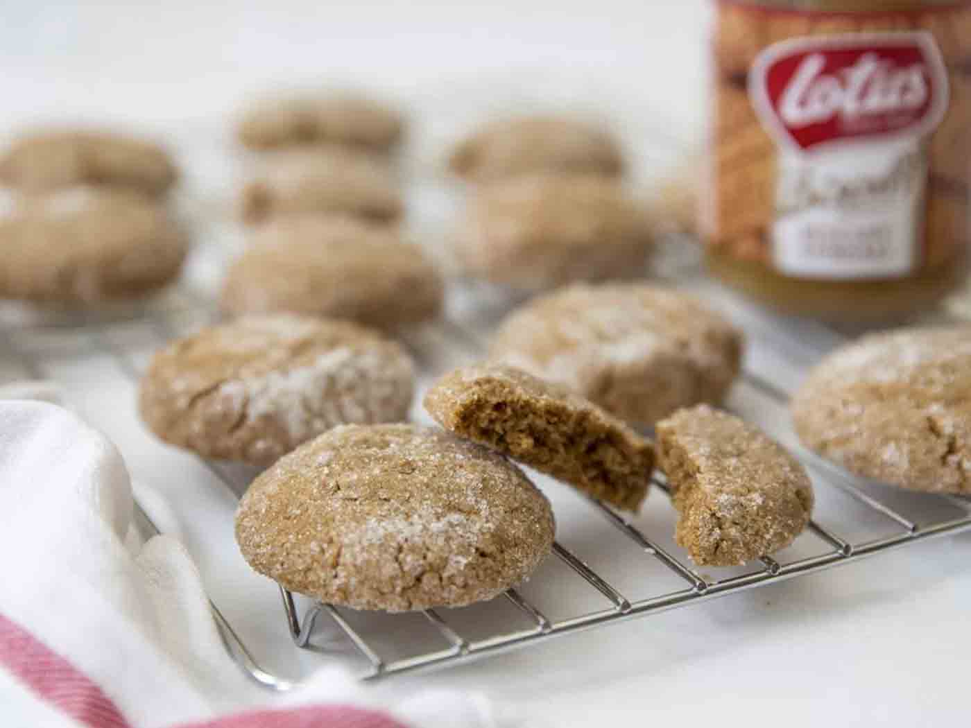 Biscoff cookies on a cooling rack. One is broken and there is a tee-towel and a jar of Biscoff in the background.
