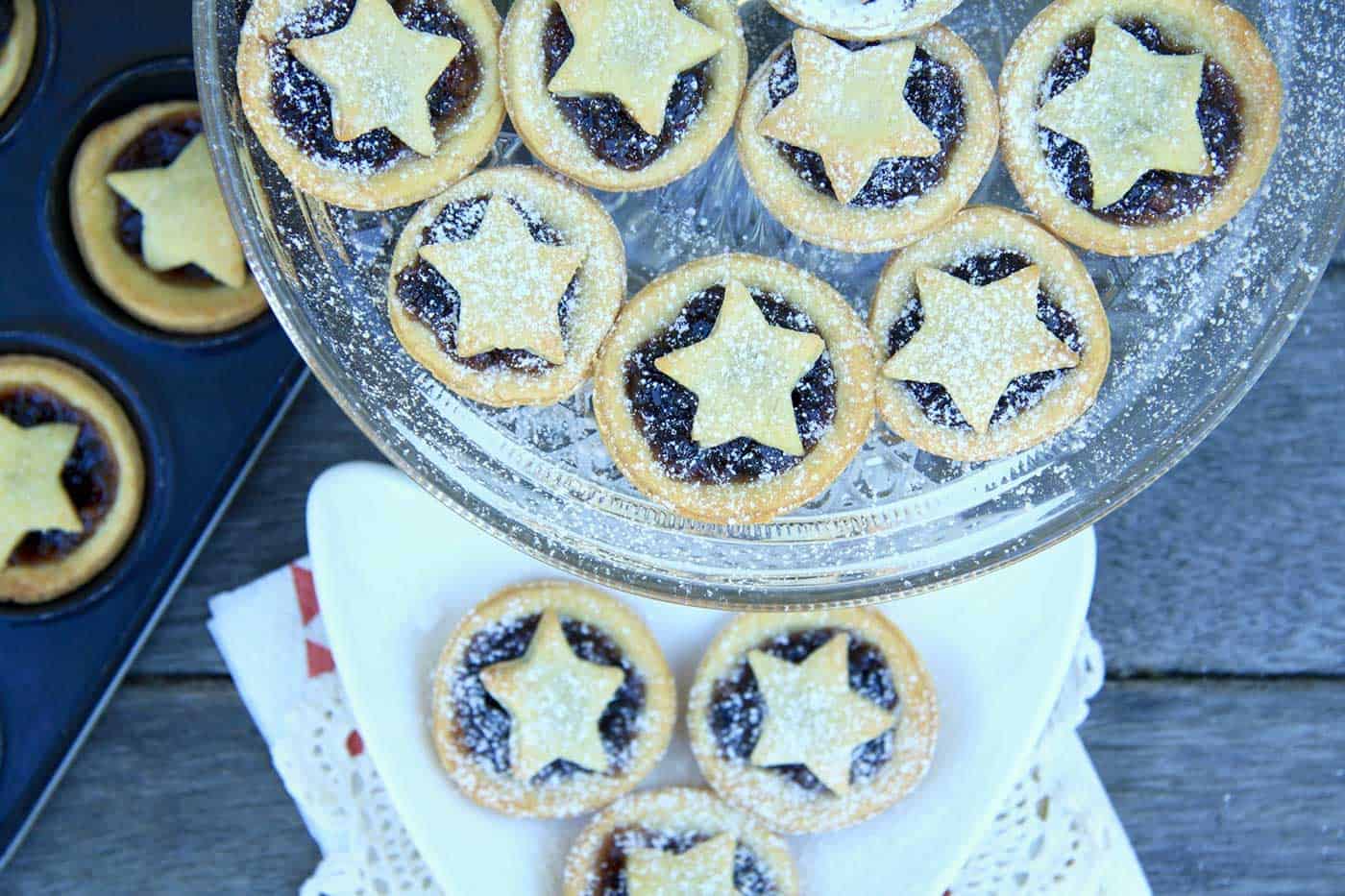 Star topped Christmas mince pies on a cake stand and sprinkled with icing sugar. More pies in the muffin tin in the background.