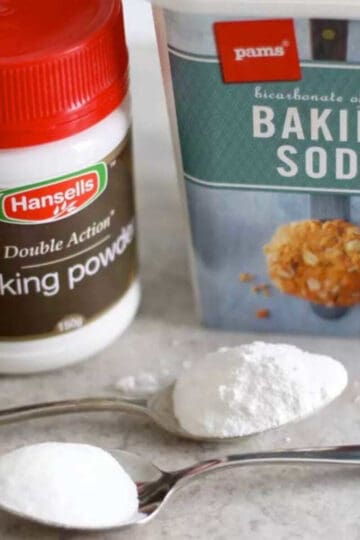 A teaspoon of baking soda and a teaspoon of baking powder. The containers of each are behind the spoons.