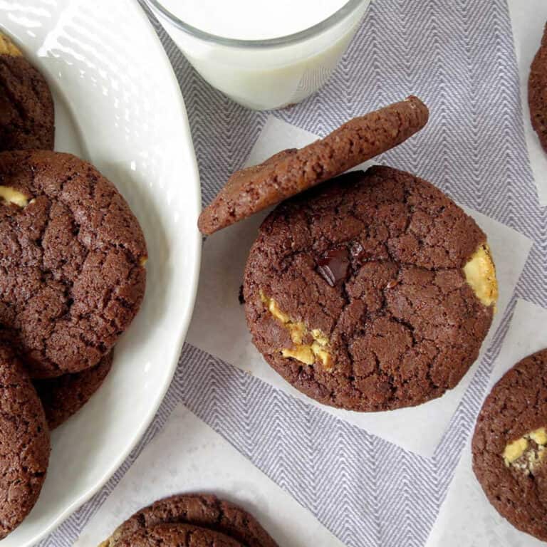 Triple chocolate chunk cookies on a piece of baking paper with other cookies surrounding it. A glass of milk also in the shot.