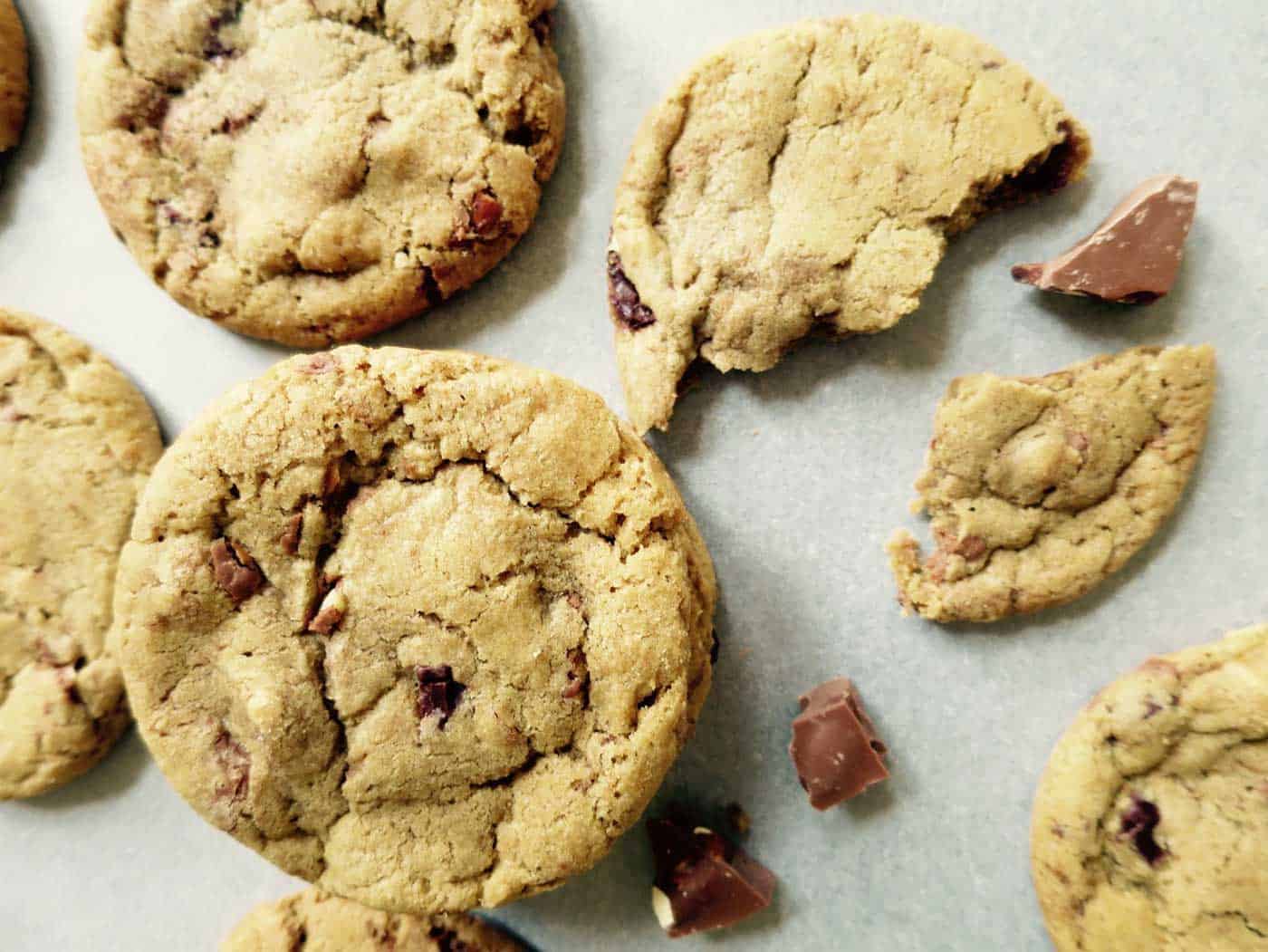 An array of chocolate chunk cookies on a tray. One is broken in half with chocolate pieces around it.