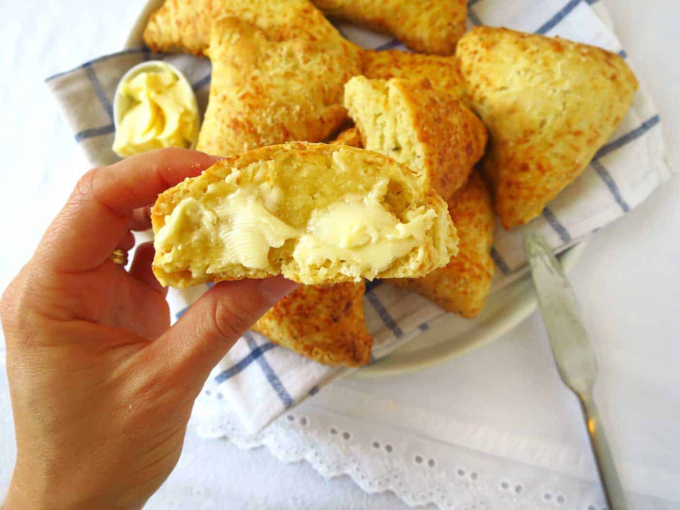 A hand holding a cheese scone lathered in butter. The remaining scones are on a plate in the background.