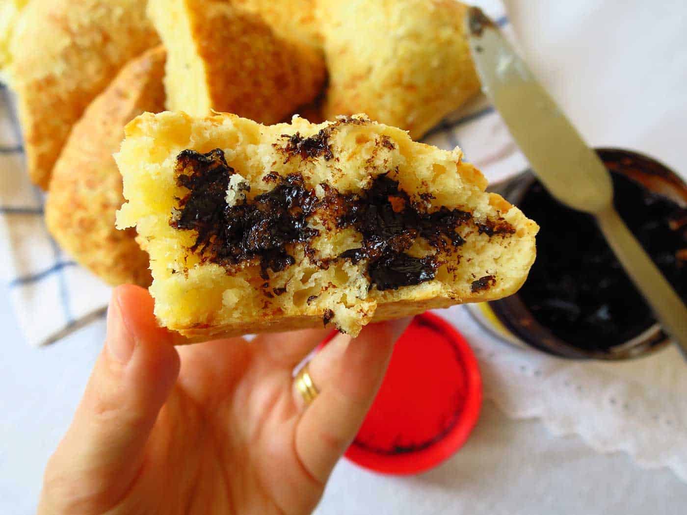 A hand holding a cheese scone lathered with butter and marmite. The marmite jar is open and a knife is resting on it.