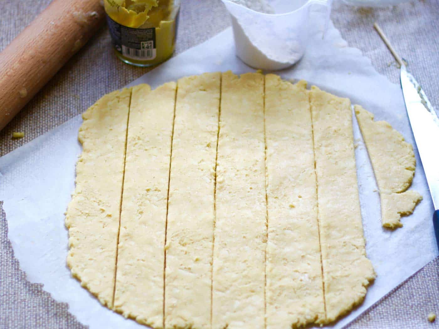 Cheese straw dough dolled out and sliced ready for baking. A rolling pin, jar of mustard and measuring cup of flour in the shot too.