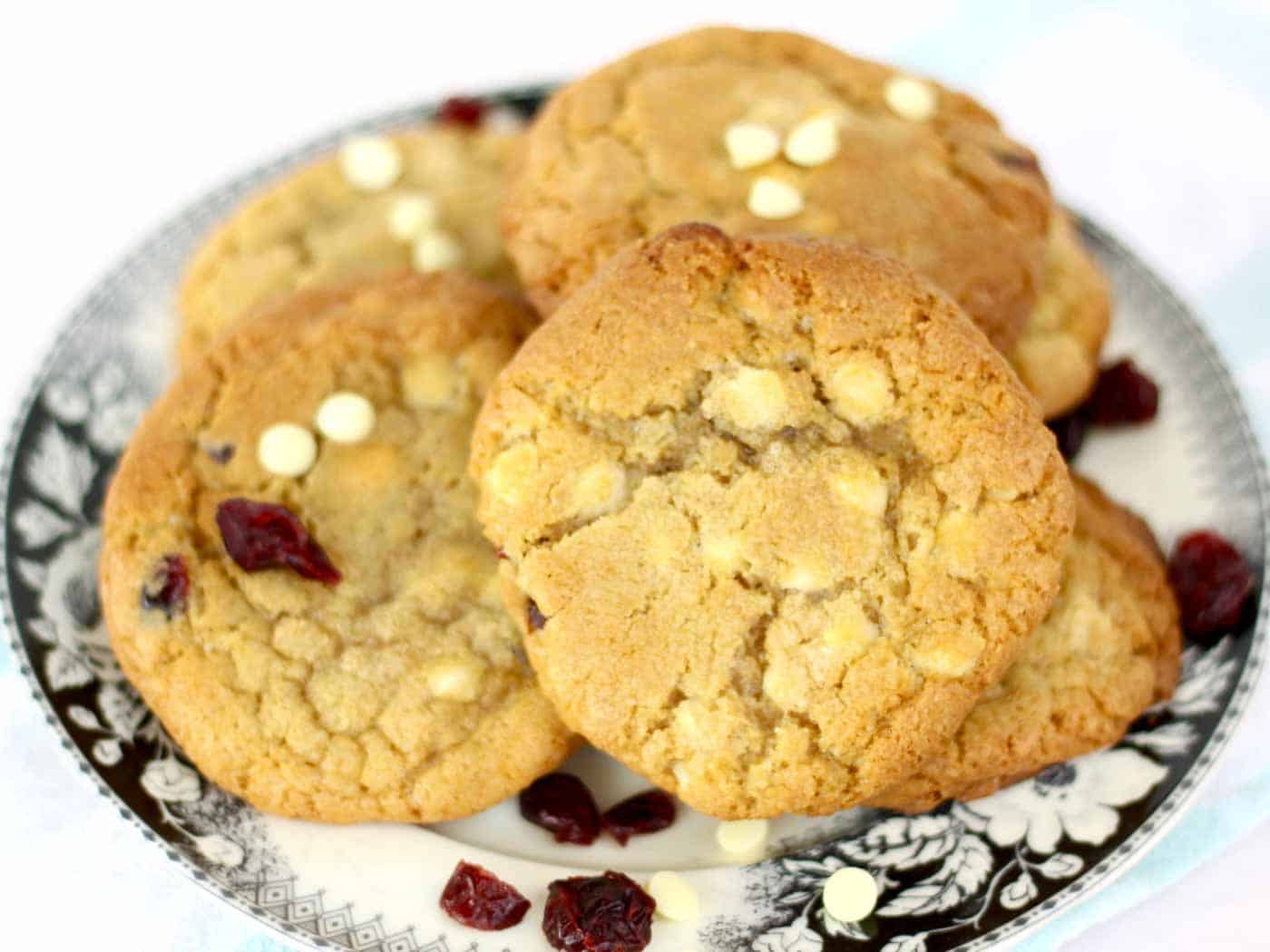 White chocolate and cranberry cookies on a plate with extra cranberries and white chocolate chips sprinkled around them.