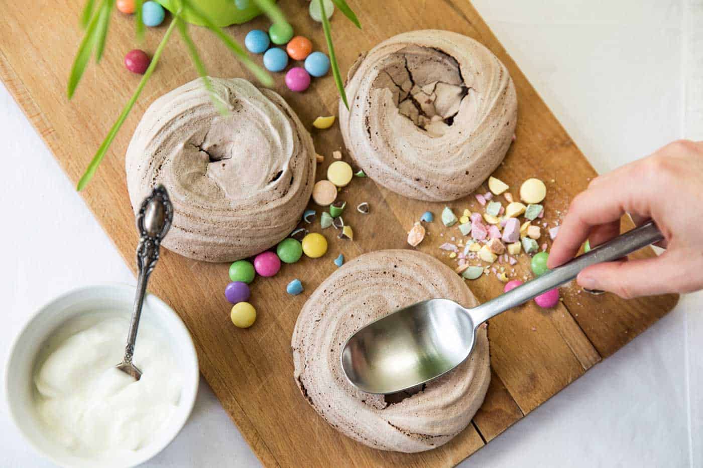 A chopping board with meringues and crushed sweets. A hand using a back of a spoon to squash meringues ready for filling. A ramekin filled with cream and a spoon.