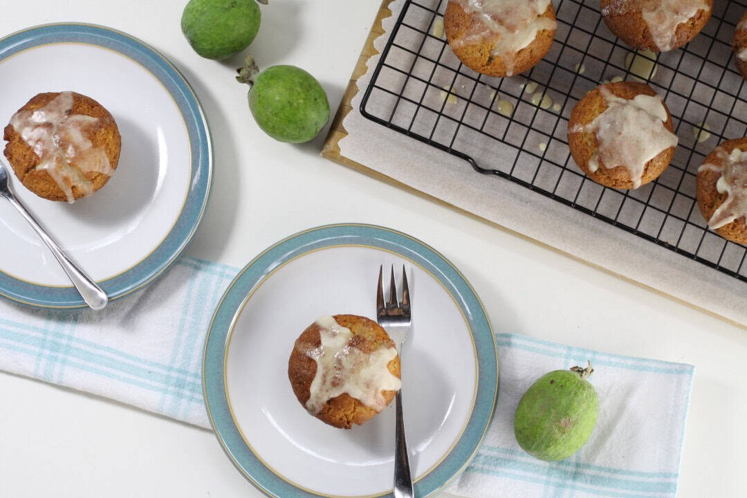 Two plates with a glazed muffin on each with forks. A cooling rack with glazed muffins and fresh feijoas in the background.