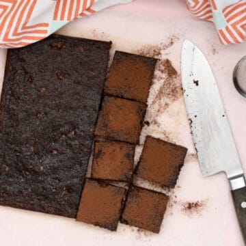 A batch of healthy chocolate brownies o na pink background. Half the batch has been sprinkled with cocoa powder and cut into squares.