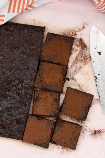 A batch of healthy chocolate brownies o na pink background. Half the batch has been sprinkled with cocoa powder and cut into squares.