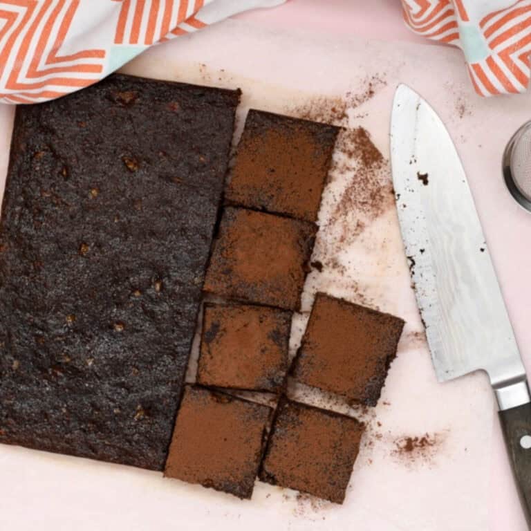 A batch of healthy chocolate brownies o na pink background. Half the batch has been sprinkled with cocoa powder and cut into squares.