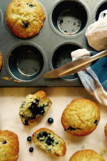 Beautiful Cafe style muffins in a muffin tin. Some muffins have been taken out of the tin and have been cut in half, showcasing lots of blueberries. A knife and t-towel are on the tin.