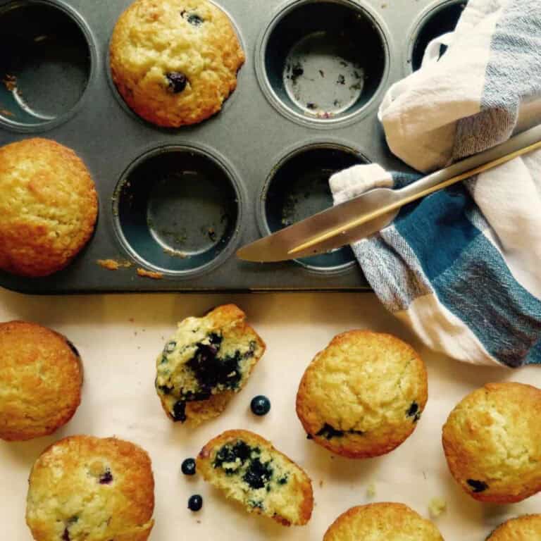 Beautiful Cafe style muffins in a muffin tin. Some muffins have been taken out of the tin and have been cut in half, showcasing lots of blueberries. A knife and t-towel are on the tin.