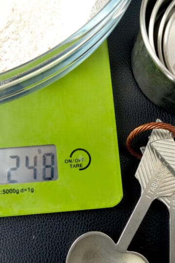Green digital scales with a bowl of flour on top. Next to it are measuring cups and measuring spoons.