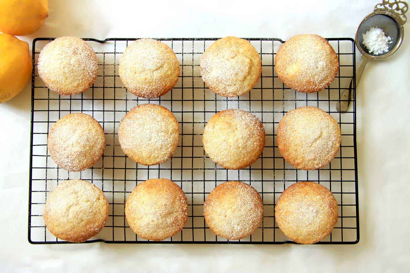 12 lemon muffins on a cooling rack sprinkled with icing sugar. Two lemons on top left corner.
