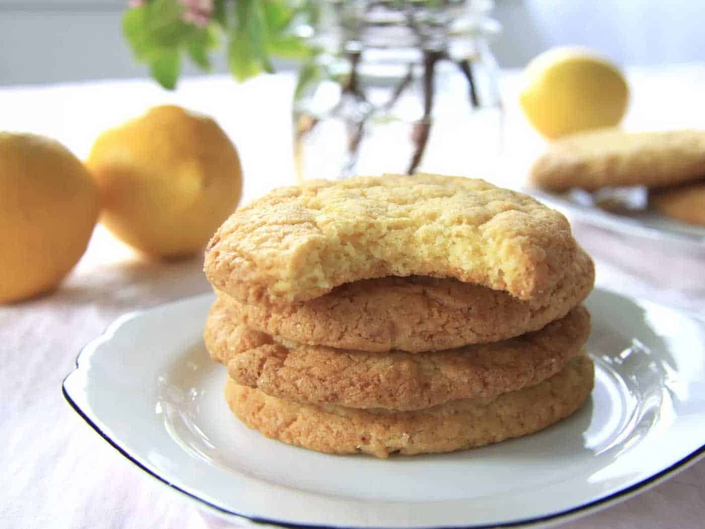 Cookies stacked on a plate with a but taken out of it. Lemons and flowers in background.