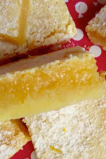 Gooey lemon bars on a white and red polkadot background. One is on it's side showcasing the flaky shortbread layer and the gooey lemon topping.