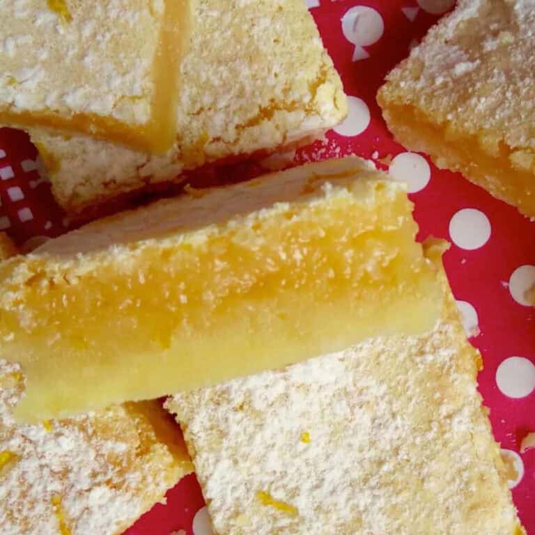 Gooey lemon bars on a white and red polkadot background. One is on it's side showcasing the flaky shortbread layer and the gooey lemon topping.