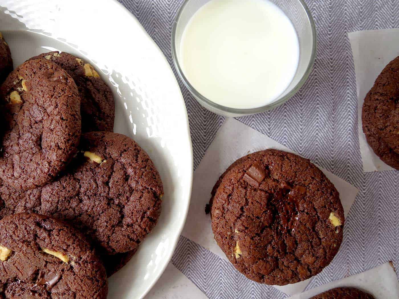 A plate of triple chocolate chunk cookies with a glass of milk and extra cookies next to it.