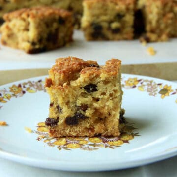 A piece of fluffy Sultana Cake on a plate iwth the remaining cake in the background. The dried fruit is distributed perfectly throughout the cake.