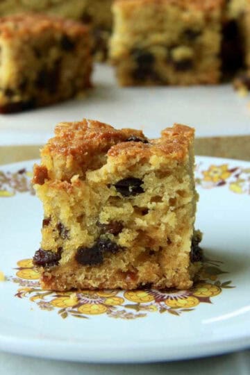 A piece of fluffy Sultana Cake on a plate iwth the remaining cake in the background. The dried fruit is distributed perfectly throughout the cake.