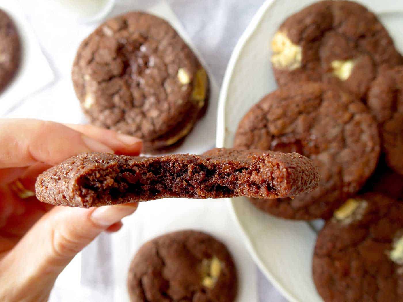 A hand holding a triple chocolate chunk cookie with a bite taken out of it and extra cookies on a plate in the background.