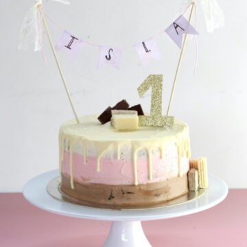 A Neapolitan cake decorated with chocolate, pink and white icing (ombre style). It has white chocolate drizzle on top with a sparkly gold number 1 topper and "Isla" cake topper.