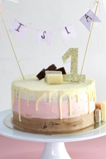 A Neapolitan cake decorated with chocolate, pink and white icing (ombre style). It has white chocolate drizzle on top with a sparkly gold number 1 topper and "Isla" cake topper.