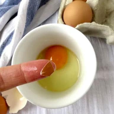 A finger with a piece of egg shell on it and a ramekin with the egg yolk and white inside.
