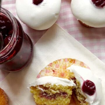 Two almond cupcakes sit on a pink and white checkered table cloth. They both have white royal icing that looks like snow with a glazed cherry on top. Next to them is a jar of jam and another almond cupcake which has been cut in half revealing the jam filling.