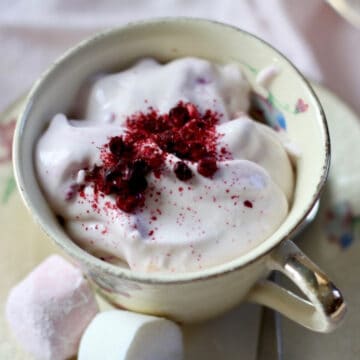 A creamy yellow coloured tea cup with fluffy pink ambrosia dessert sprinkled with berry powder. Two marshmallows are on the side of the tea cup on the saucer.
