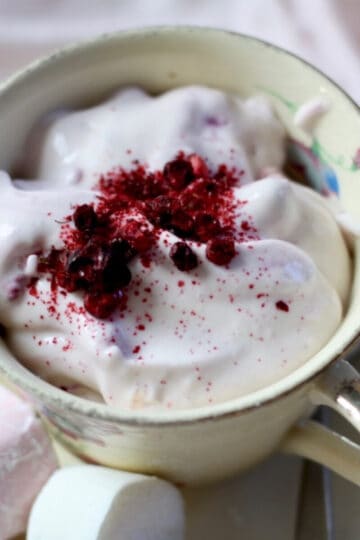 A creamy yellow coloured tea cup with fluffy pink ambrosia dessert sprinkled with berry powder. Two marshmallows are on the side of the tea cup on the saucer.