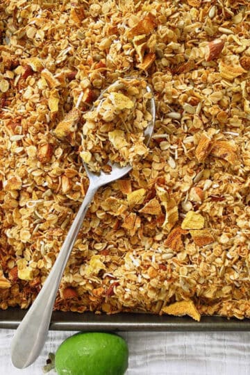 A baking tray with feijoa granola on it with a serving spoon on it ready to scoop into bowls. There are several fresh feijoas on the side of the baking tray.