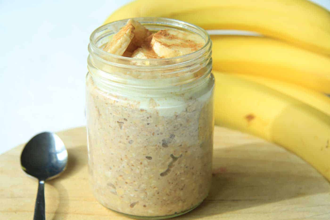 A glass jar filled with banana bread overnight oats. Bananas in the background.