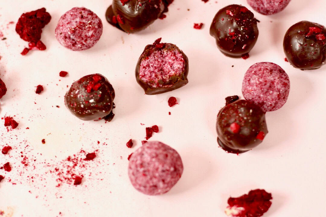 Berry bliss balls on a pink board. Some are coated in chocolate with extra berry dust sprinkled on top and others are left plain without chocolate. The pink berries are vibrant!