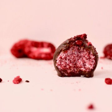 A pink background with a berry explosion bliss ball that has been cut in half. The inside of the bliss ball is a bright pink colour with specks of coconut through it. The casing of the chocolate shell is thick and looks so delicious. There are freeze dried berries in the background.
