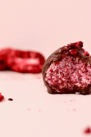 A pink background with a berry explosion bliss ball that has been cut in half. The inside of the bliss ball is a bright pink colour with specks of coconut through it. The casing of the chocolate shell is thick and looks so delicious. There are freeze dried berries in the background.