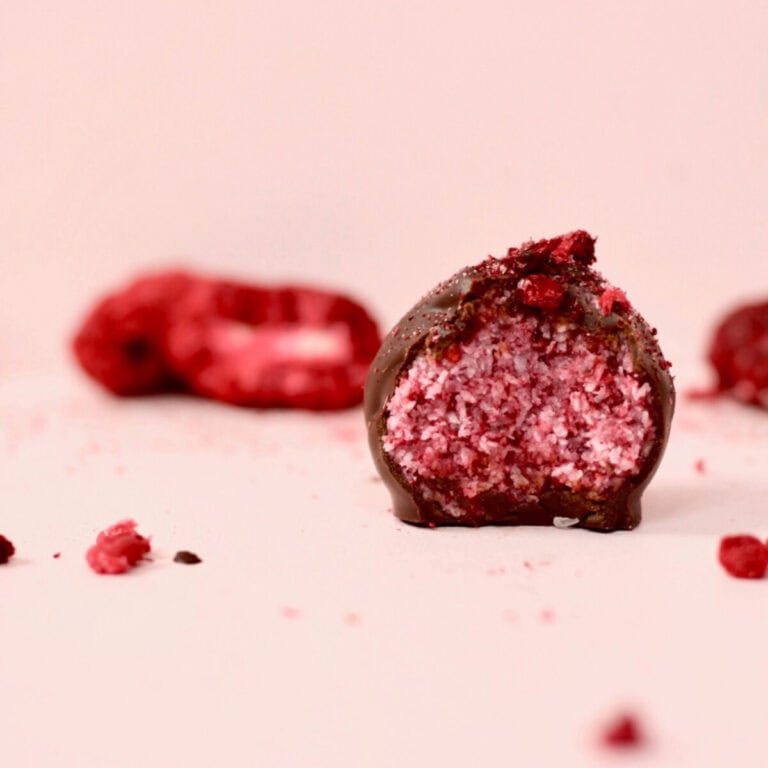 A pink background with a berry explosion bliss ball that has been cut in half. The inside of the bliss ball is a bright pink colour with specks of coconut through it. The casing of the chocolate shell is thick and looks so delicious. There are freeze dried berries in the background.