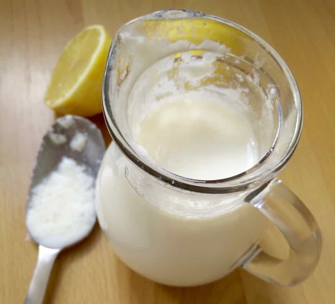 A jug filled with buttermilk. A stiring spoon with buttermilk on it next the jug and lemon halves also.