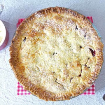 A fully baked cherry pie with perfectly crimped edges sitting on a red and white checkered tea towel. There is a ramekin with cream next to it and a cake server ready to serve the first slice.
