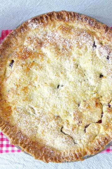 A fully baked cherry pie with perfectly crimped edges sitting on a red and white checkered tea towel. There is a ramekin with cream next to it and a cake server ready to serve the first slice.