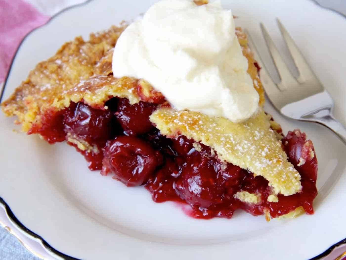 A close up shot of a delectable slice of cherry pie, with cherries oozing out the sides. A dollop of ice cream sits on top of the pie slice.