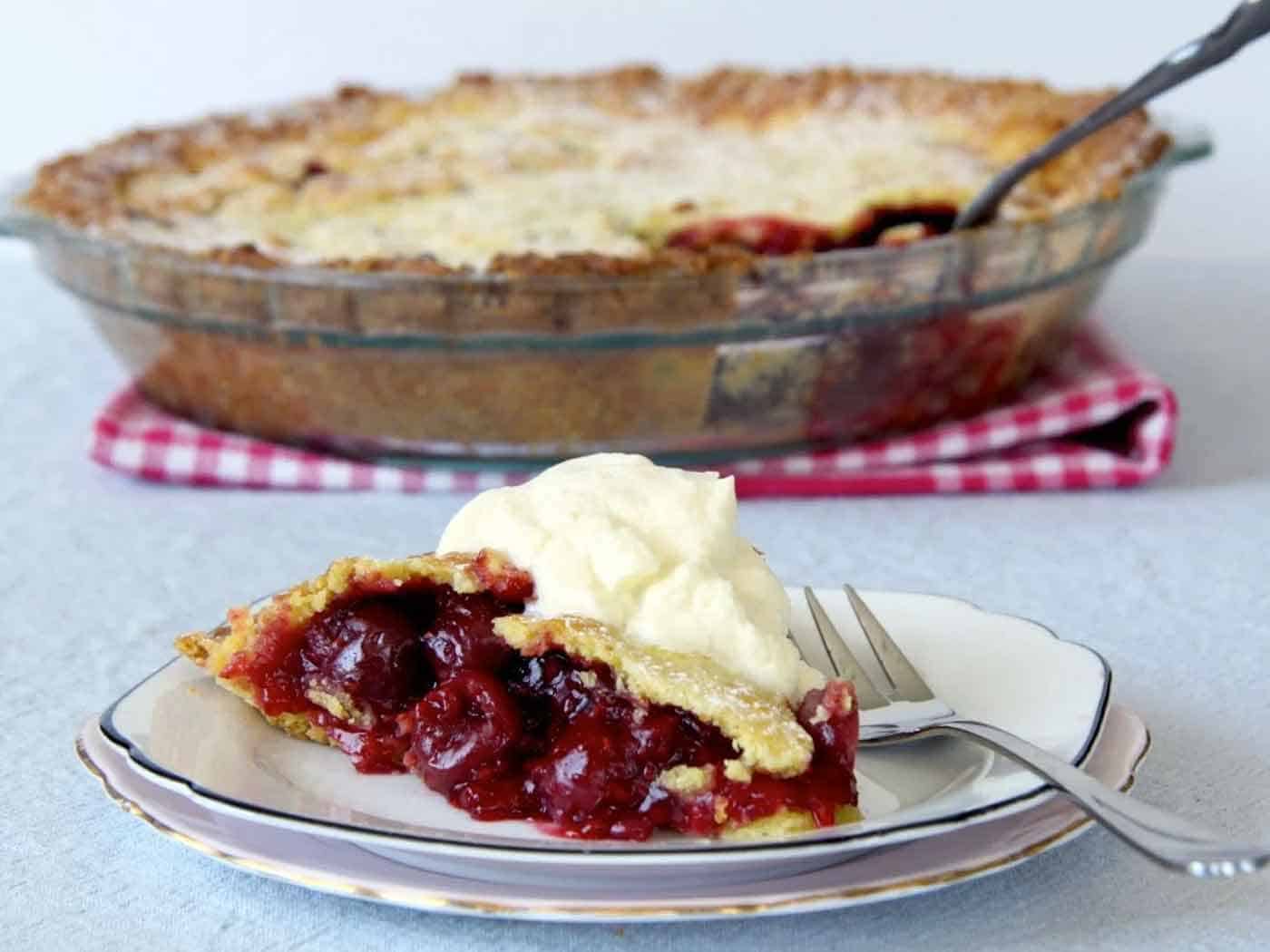 A slice of cherry pie with ice cream on the side on a plate. The rest of the pie in the background, still in the pie dish.