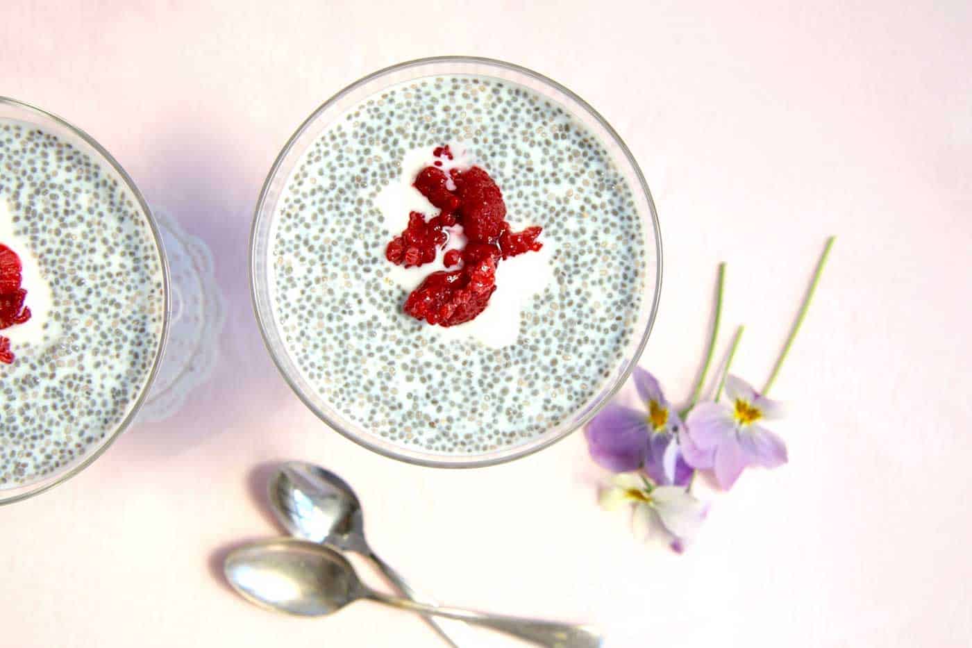 Birds-eye view of chia seed pudding. Each are topped with a dollop of yoghurt and cooked berries. Teaspoons and violet flowers used for decoration on the side.