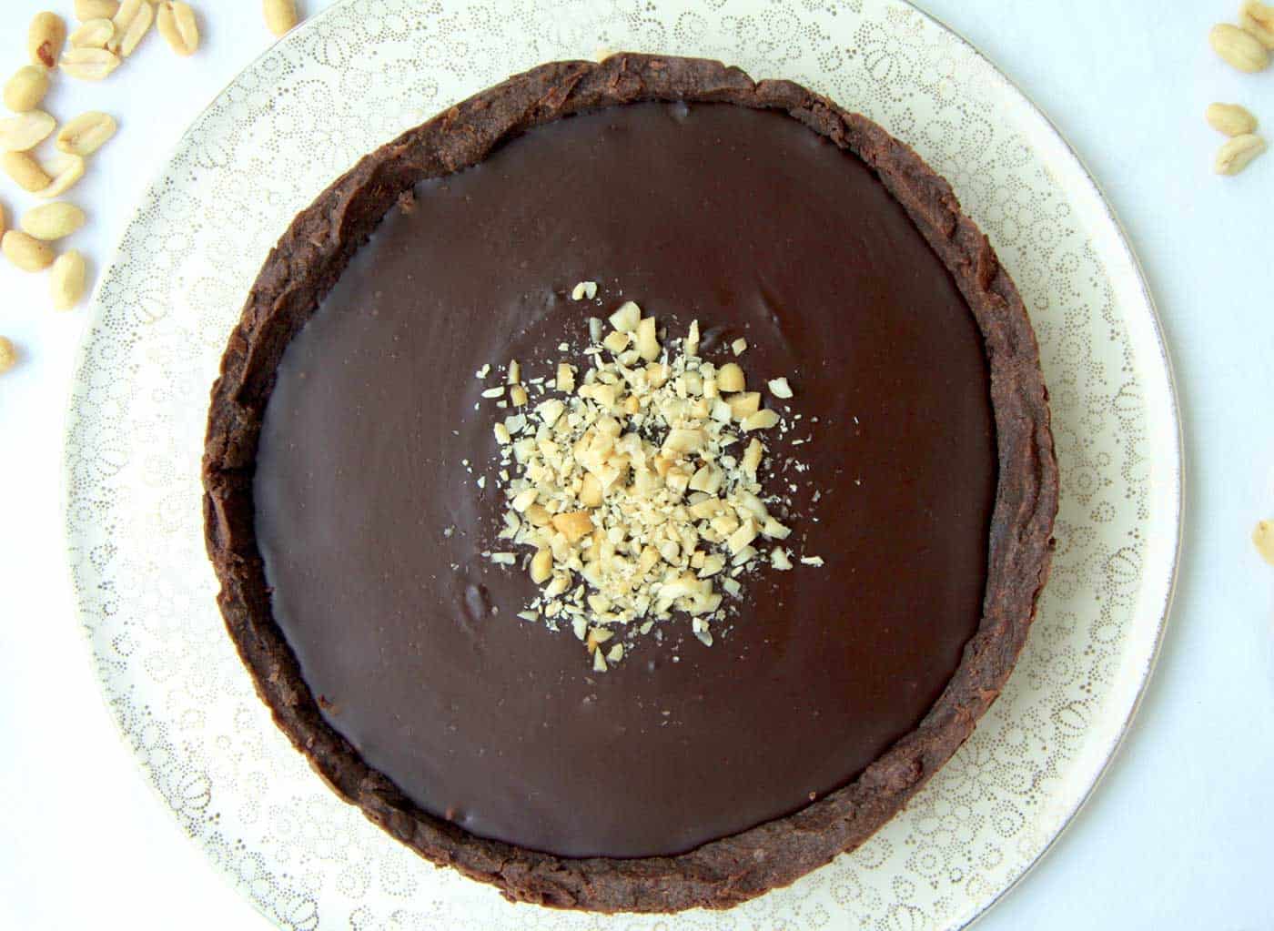 A chocolate tart on a serving plate before being cut into. Chopped nuts are sprinkled in the middle of the tart with extra nuts on the side. White table cloth background.