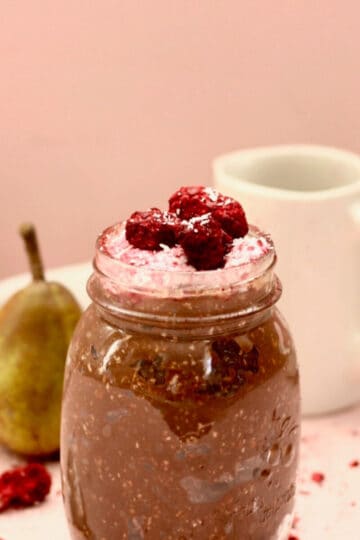 A jar filled with chocolate coconut bircher muesli with berry yoghurt on top and extra berries to decorate. There is a pink backdrop , two rustic green pears and a small white milk jug next to it.