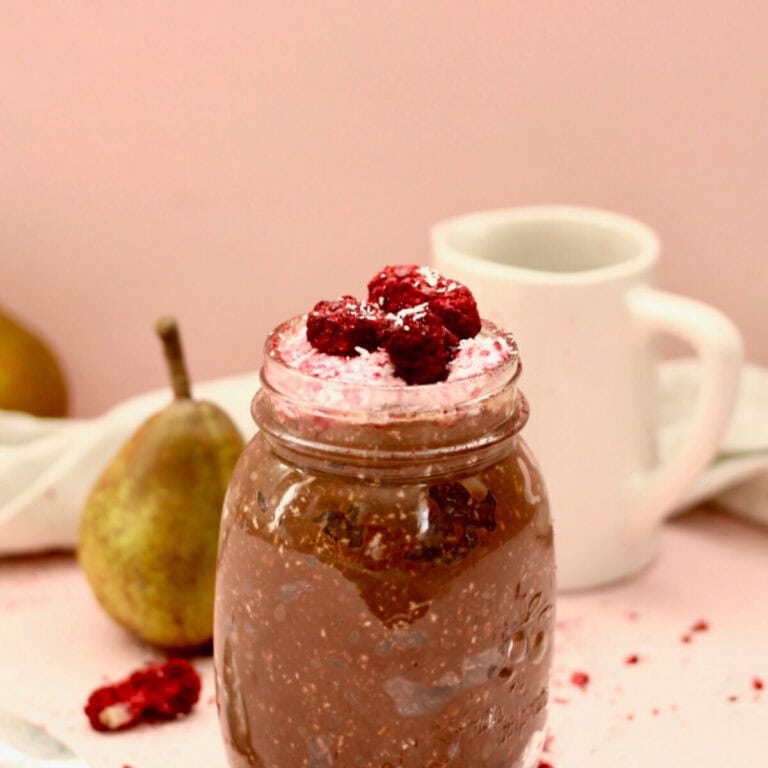 A jar filled with chocolate coconut bircher muesli with berry yoghurt on top and extra berries to decorate. There is a pink backdrop , two rustic green pears and a small white milk jug next to it.