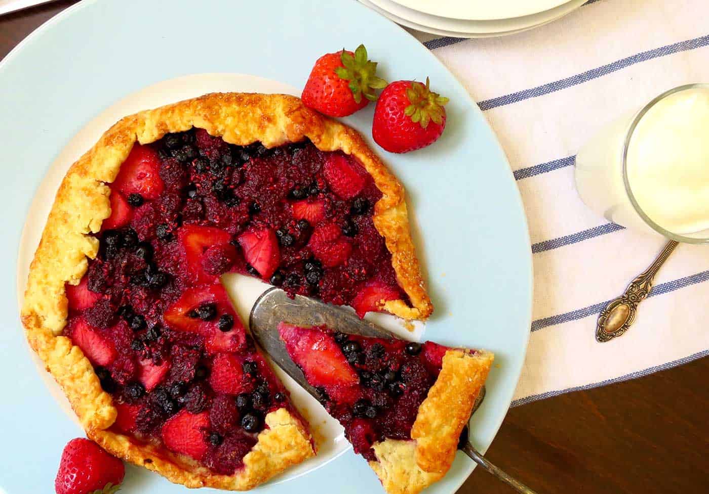 A top view of the berry galette with a cake server about to serve a slice of galette on to a side plate. Yoghurt in a jar on the side.
