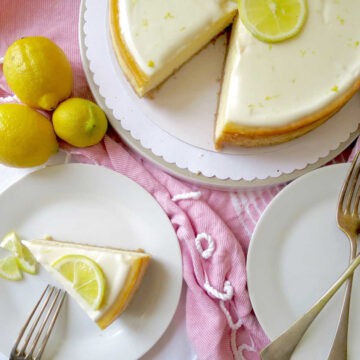 A velvety smooth and luscious looking lemon cheesecake on a pink table cloth. A slice of cheese cake is next to it on a side plate and there are fresh lemons beside it.